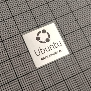 May include: A square, silver sticker with the Ubuntu logo, featuring three connected circles above the word "Ubuntu" and the text "open source". The sticker is on a gray grid background.