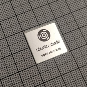 Peut inclure: Un autocollant carré argenté avec les mots "ubuntu studio" et "open source" en police sans-serif. L'autocollant présente un logo circulaire noir avec des cercles concentriques. L'autocollant est sur un fond de grille grise.