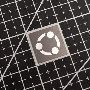 Könnte beinhalten: Ein quadratischer Aufkleber mit grauem Hintergrund und einem weißen kreisförmigen Logo, das aus drei verbundenen Kreisen besteht. Der Aufkleber befindet sich auf einer schwarzen Schneidematte mit weißen Gitterlinien.