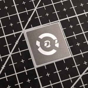 Pode incluir: Um autocolante quadrado cinzento com um design branco com um símbolo "@" estilizado dentro de um padrão circular. O autocolante está sobre uma base de corte preta com linhas de grelha brancas.