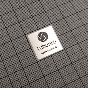 Peut inclure: Un autocollant carré argenté avec le logo Lubuntu, représentant un oiseau stylisé dans un cercle, et les mots "Lubuntu open source" en police sans-serif. L'autocollant est sur un fond quadrillé gris.
