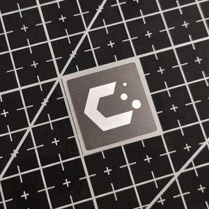 May include: A square sticker with a grey background and a white geometric design. The design features a stylized letter "C" and two white circles. The sticker is placed on a black cutting mat with white grid lines.
