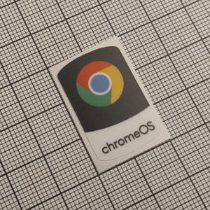 May include: A rectangular sticker with the ChromeOS logo, featuring a colorful circular design in red, yellow, green, and blue, set against a dark gray background. The text "chromeOS" is printed below the logo.