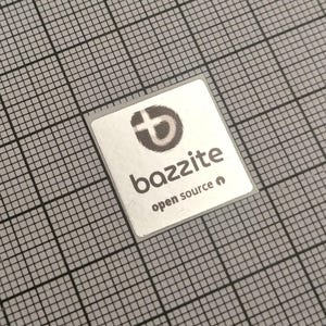 Op de afbeelding: Een kleine, vierkante sticker met het woord "bazzite" in een modern lettertype en de tekst "open source" eronder. De sticker heeft een zilveren achtergrond met een zwart-grijs rasterpatroon op de achtergrond. Het "bazzite"-logo is een gestileerde "b" met een kruis.