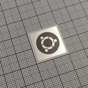 May include: A square sticker with a silver background and a black Ubuntu logo. The logo features a central circle surrounded by three smaller circles. The sticker is placed on a gray grid background.