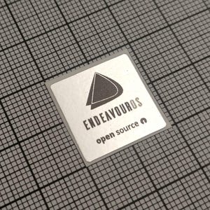 May include: A square silver sticker with the word "ENDEAVOUROS" in a sans-serif font, and the words "open source" below it. The sticker has a dark gray triangle logo. The sticker is on a gray grid background.
