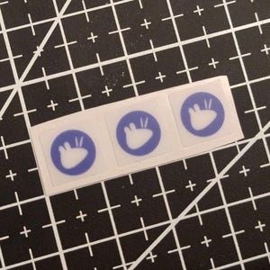 Op de afbeelding: Drie vierkante stickers in een horizontale rij, elk met een cirkelvormige blauwe achtergrond en een witte afbeelding van een gestileerde konijnenkop met twee rechtopstaande oren. De stickers zitten op een wit vel.