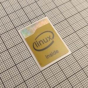 Pode incluir: Um autocolante dourado com o texto "(linux) inside" em preto. O autocolante tem a forma de um retângulo arredondado com uma borda branca e está colocado sobre um fundo quadriculado. A parte superior do autocolante tem um efeito holográfico.