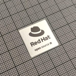 Può includere: Un adesivo quadrato argentato con le parole "Red Hat" in grassetto, grigio scuro, e le parole "open source" sotto. Un'icona di cappello fedora grigio scuro è sopra il testo. L'adesivo è su uno sfondo a griglia grigia.
