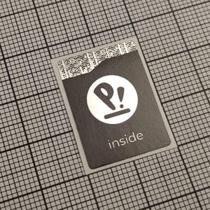 Pode incluir: Um autocolante retangular preto com um logótipo branco e a palavra "inside" impressa em branco. O logótipo é um círculo branco com um "P" estilizado e um ponto de exclamação. Fundo de grelha cinzenta.