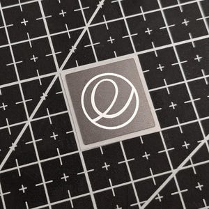 Op de afbeelding: Een vierkante, grijze sticker met een wit, gestileerd letter "e" ontwerp. De sticker ligt op een zwarte snijmat met witte rasterlijnen en plustekens.
