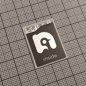 May include: A rectangular black and silver sticker with the word "inside" printed in white. The sticker features a white graphic of a bird-like shape. The background is a gray grid.
