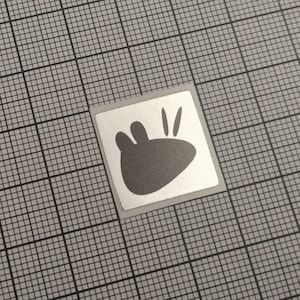 Könnte beinhalten: Ein kleiner, quadratischer Aufkleber mit einer grauen Grafik eines stilisierten Pfotenabdrucks mit drei Zehen und zwei Linien. Der Aufkleber hat einen silbernen Rand und befindet sich auf einem grauen Gitterhintergrund.