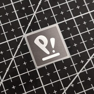 Può includere: Un adesivo quadrato con sfondo grigio e un design bianco con le lettere "P!" e una linea orizzontale. L'adesivo è su un tappetino da taglio nero con linee a griglia bianche e segni più.