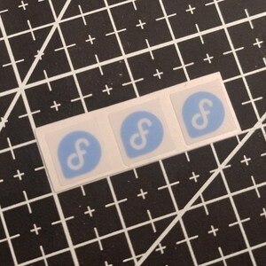 Op de afbeelding: Drie vierkante stickers in een horizontale rij. Elke sticker heeft een lichtblauw druppelvormig icoon met een witte kleine letter f. De stickers hebben een dunne witte rand en liggen op een zwarte snijmat met een rasterpatroon.