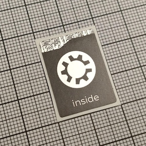 Könnte beinhalten: Ein rechteckiger Aufkleber mit schwarzem Hintergrund und einem weißen Zahnradsymbol in einem weißen Kreis. Das Wort "inside" ist darunter in Weiß gedruckt. Der Aufkleber befindet sich auf einem grauen Raster.