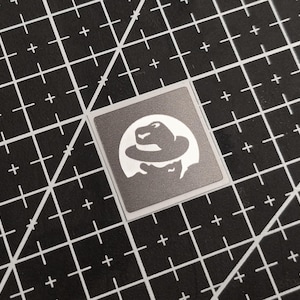 May include: Square sticker with a silver background and a white graphic of a person wearing a fedora hat. The sticker is on a black cutting mat with white grid lines.