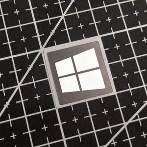 Könnte beinhalten: Ein quadratischer Aufkleber mit dem Windows-Logo, einem weißen Quadrat, das in vier Teile geteilt ist, auf dunklem Hintergrund. Der Aufkleber liegt auf einer schwarzen Schneidematte mit weißen Rasterlinien.