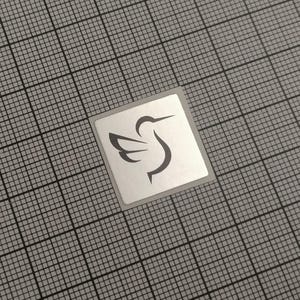 Könnte beinhalten: Ein quadratischer Aufkleber mit silbernem Rand und einem schwarzen Kolibri-Design. Der Kolibri ist in einem minimalistischen Stil dargestellt, mit ausgebreiteten Flügeln. Der Aufkleber befindet sich auf einem grauen Gitterhintergrund.