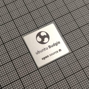 May include: A square, silver sticker with the text "ubuntu Budgie" and "open source" in a sans-serif font. The sticker features a black and white logo of three connected shapes. The sticker is on a gray grid.