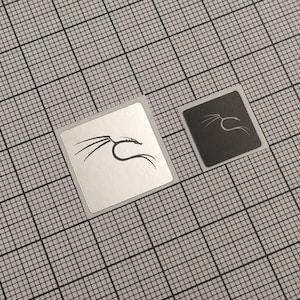 Peut inclure: Deux autocollants carrés avec un motif de dragon stylisé. L'un est blanc avec un dragon noir, l'autre est noir avec un dragon blanc. Ils sont posés sur un fond de grille grise.