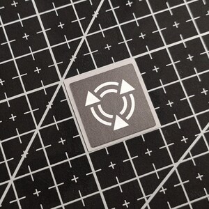 Könnte beinhalten: Ein quadratischer, grauer Aufkleber mit einem weißen, kreisförmigen Pfeilsymbol. Der Aufkleber befindet sich auf einer schwarzen Schneidematte mit einem Rastermuster.