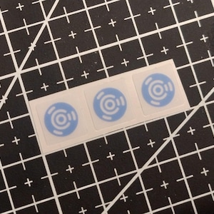 Op de afbeelding: Drie vierkante stickers in een horizontale rij, elk met een lichtblauw cirkelvormig ontwerp met een centrale stip omringd door twee concentrische ringen en twee gebogen segmenten, gedrukt op transparant zelfklevend materiaal.