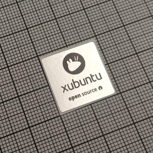 Pode incluir: Um autocolante quadrado prateado com a palavra "xubuntu" em fonte sem serifa e as palavras "open source" por baixo. O autocolante tem um logótipo circular preto com forma de mão. O autocolante está numa grelha cinzenta.