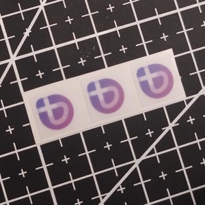 May include: Three square stickers arranged in a horizontal row, each featuring a circular purple and white logo with a stylized letter b and a cross symbol. The stickers are placed on a black cutting mat with a white grid of lines and plus signs.