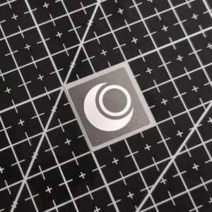 Peut inclure: Un autocollant carré avec une bordure grise et un motif blanc de croissant de lune et de cercle. L'autocollant est sur un tapis de coupe noir avec des lignes de grille blanches et des signes plus.