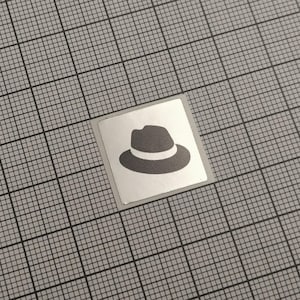 Może przedstawiać: Kwadratowa naklejka z ciemnoszarym kapeluszem fedora z białą opaską. Naklejka znajduje się na szarym tle w kratkę.