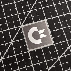 Könnte beinhalten: Quadratischer Aufkleber mit weißem Commodore-Logo auf grauem Hintergrund. Der Aufkleber befindet sich auf einer schwarzen Schneidematte mit weißen Rasterlinien und Pluszeichen.