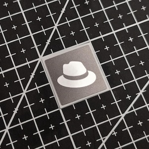 May include: A square sticker with a white fedora hat design on a gray background. The sticker is set against a black cutting mat with white grid lines and plus signs.