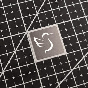 May include: A square sticker featuring a white hummingbird design on a grey background. The sticker is on a black cutting mat with white grid lines and plus signs.