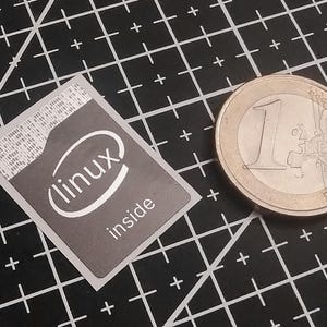 May include: A rectangular sticker featuring a dark gray background with the white text "linux inside" and a stylized oval graphic. The sticker is positioned next to a one-euro coin for scale, which has a diameter of approximately 23.25 millimeters.