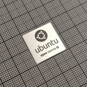 May include: A square, silver sticker with the Ubuntu logo, a circle with three connected dots above a smaller circle. The word "ubuntu" is below the logo, with "open source" beneath it. The sticker is on a gray grid.
