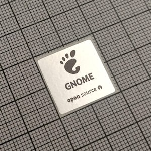 Op de afbeelding: Een vierkante, zilveren sticker met het woord "GNOME" in zwart, een pootafdruklogo en de tekst "open source". De sticker bevindt zich op een grijze rasterachtergrond.