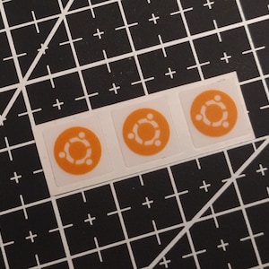 Op de afbeelding: Drie vierkante stickers in een horizontale rij, elk met een cirkelvormig oranje icoon met in het midden een gestileerde witte afbeelding van drie personen. De stickers liggen op een zwarte snijmat met een wit raster van lijnen en plusjes.