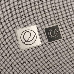May include: Two square stickers with a black line drawing of a stylized letter "e" on a grid background. One sticker is silver, the other is black. The "e" design is a continuous line with a loop.