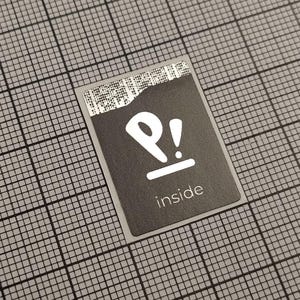 Pode incluir: Um autocolante retangular preto com um logótipo branco e a palavra "inside". O logótipo é um "P" estilizado com um ponto de exclamação e uma linha horizontal por baixo. A extremidade superior tem um código de barras.