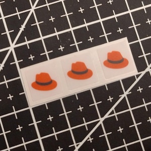 May include: A rectangular sticker sheet featuring three individual square stickers, each displaying a graphic illustration of an orange fedora hat with a dark gray band. The stickers are arranged in a horizontal row on a white backing.