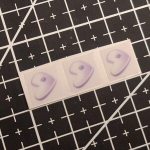 May include: Three small, square stickers arranged in a horizontal row. Each sticker features a light purple, comma-shaped graphic with a small circular dot near the top curve, set against a white background on the adhesive backing.
