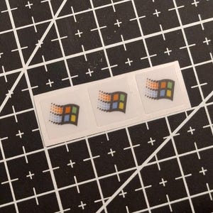 Puede incluir: Una tira de tres pegatinas cuadradas pequeñas, cada una con un diseño clásico de logotipo de ventana ondulante multicolor con paneles rojos, verdes, azules y amarillos sobre un fondo blanco, dispuestas horizontalmente en una fila.
