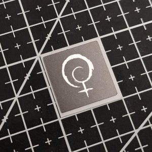 Op de afbeelding: Een vierkante sticker met een wit symbool van een cirkel met een spiraal en een kruis eronder, op een grijze achtergrond. De sticker ligt op een zwarte snijmat met witte rasterlijnen en plustekens.