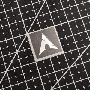 May include: A square sticker featuring the Arch Linux logo, a stylized white 'A' shape on a dark gray background. The sticker is set against a black cutting mat with white grid lines and diagonal markings.