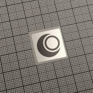 May include: A small, square sticker with a silver border. The sticker features a dark grey design of a crescent moon shape with a circle inside, and another circle around the outside. The sticker is on a grey grid background.