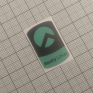 May include: A rectangular sticker with a black background and a teal-colored circle and arrow design. The bottom of the sticker has the words "Rocky Linux" in teal. The sticker is on a grid background.