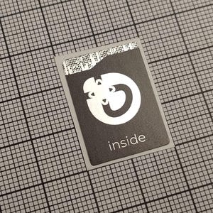 Könnte beinhalten: Ein rechteckiger schwarzer Aufkleber mit einem weißen Logo und dem Wort "inside" in Weiß gedruckt. Das Logo ist ein stilisiertes Design mit einer kreisförmigen Form und zwei gebogenen Elementen. Der Aufkleber hat einen hellgrauen Rand.
