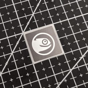 May include: A square, grey sticker featuring a white eye graphic inside a circle. The sticker is placed on a black cutting mat with white grid lines. The eye design is a stylized illustration.