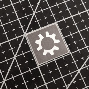 Könnte beinhalten: Ein quadratischer Aufkleber mit grauem Hintergrund und einem weißen Zahnradsymbol. Der Aufkleber befindet sich auf einer schwarzen Schneidematte mit weißen Gitterlinien und Pluszeichen.